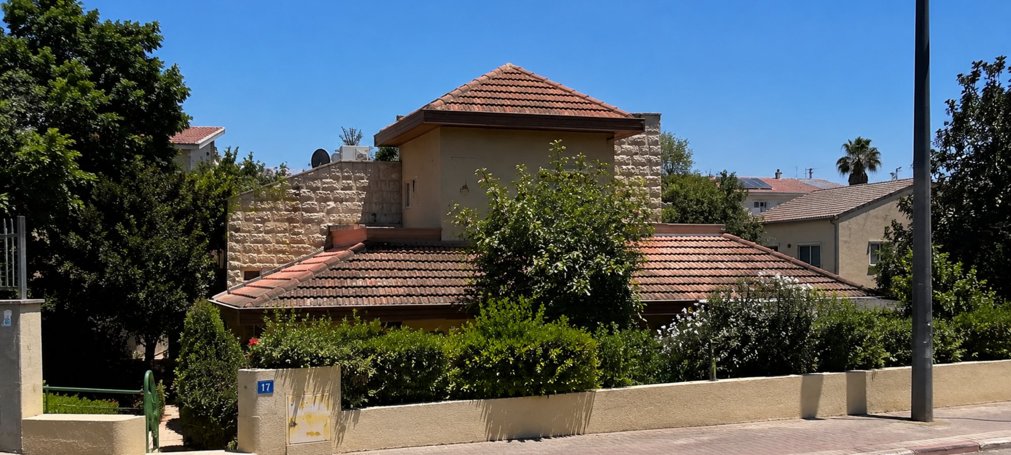 A stone-clad two-storey Israeli house behind a rendered boundary wall and clipped hedge in bright midday sun