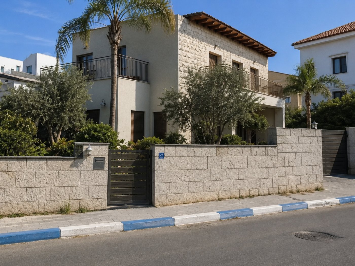 A two-storey Israeli house in Jerusalem stone and pale render, set behind a stone boundary wall with a gate, olive trees and palms